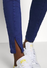 Blå denimjeans med sidöppningar vid fållen, i kombination med vita sneakers med en gul accent och klassisk snörningsdesign.