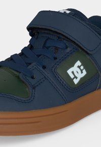 Baskets enfant bleu marine et vert avec semelle marron, lacets et bande Velcro, avec un logo DC blanc sur le côté.