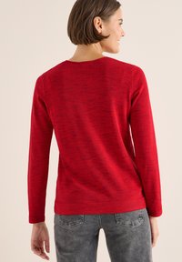 Maglione rosso a maniche lunghe con un design di maglia testurizzata e un classico collo rotondo, abbinato a jeans grigi e una vestibilità leggermente rilassata.
