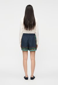 Calções azul-marinho com bolinhas brancas, cintura elástica e detalhes às riscas multicoloridas na bainha. Modelo a usar uma blusa creme de manga comprida.