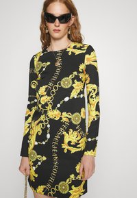 Vestido negro de manga larga con un atrevido patrón barroco amarillo y acentos dorados. El diseño incluye botones blancos y texto de logo en todo el vestido.