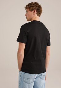 Camiseta de algodón negra con cuello redondo y mangas cortas, vista desde la parte de atrás, combinada con jeans de mezclilla azul claro.