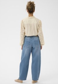 Kremhvid bluse med blonder på ærmerne, kombineret med lyseblå, løstsiddende jeans og beige sko. Blusen har krave med flæser og detaljer med fine læg.