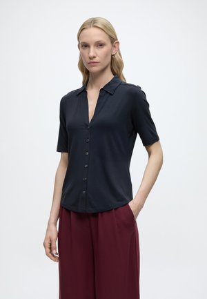 Blonde Frau mit glattem Haar trägt ein schwarzes, kurzärmeliges, geknöpftes Hemd und lockere bordeauxrote Hosen, steht mit einer Hand in der Tasche.