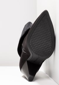 Botte en daim noir à bout pointu avec semelle en caoutchouc texturée et talon empilé de hauteur moyenne, inclinée contre une surface d'angle blanche.