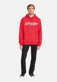 Haut à capuche rouge avec un logo "SPYDER" blanc, conçu avec une poche kangourou et des cordons de serrage réglables. Assorti à un pantalon noir et des baskets.