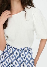 Blouse blanche texturée à manches courtes bouffantes, associée à une jupe bleue et blanche à motifs de losanges.