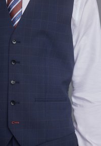 Navy kariertes Weste mit sechs Knöpfen, schwarzem Knopfdetail und rotem Akzentstich. Über einem weißen Hemd getragen, das einen formellen Stil zur Geltung bringt.