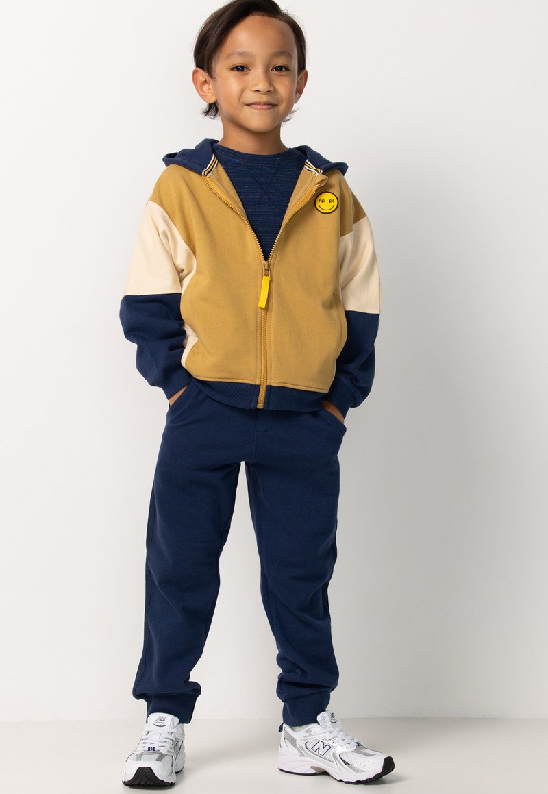 Gelb und marineblau blockierte Zip-Hoodie mit einem Smiley-Patch, kombiniert mit marineblauen Jogginghosen. Die Sneaker haben graue und schwarze Akzente.