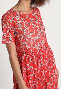 Červené květinové šaty s kulatým výstřihem, krátkými rukávy a rozšířenou sukní zdobenou černobílými květinovými vzory.