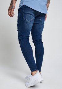 Mörkblå slim-fit jeans med en slitning på vänster baklår, ihop med vita sportskor.