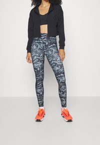 Czarna krótkia bluza z kapturem na zamek, czarny stanik sportowy, wzorzyste legginsy w odcieniach niebiesko-szarych oraz jaskrawe pomarańczowe buty do biegania z czarnymi akcentami.