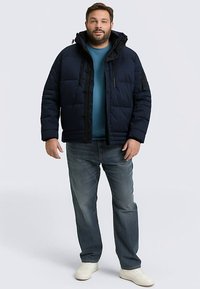 Gewatteerd marineblauw jack met capuchon, voorzien van ritsen en zakken aan de voorkant, gedragen over een teal gekleurd shirt en blauwe spijkerbroek, gecombineerd met witte sneakers.