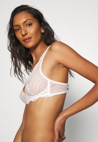 Blondet bralette i hvit med geometrisk mønster, med brede stropper, et semi-gjennomsiktig design og utklippede detaljer på sidene.
