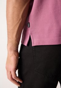 Polo shirt rose texturé avec fentes latérales, étiquette de la marque "BOSS," porté avec un pantalon en denim noir, présentant un gros plan d'une main près de l'ourlet.