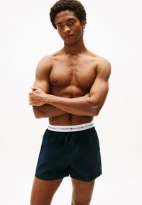 Shorts de bain bleu marine avec une ceinture élastique blanche mettant en avant le logo "TOMMY HILFIGER". Design simple et fonctionnel avec une ouverture frontale discrète.