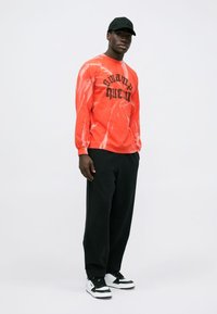 Long-sleeve, orange tie-dye shirt with "swamp queen" text, paired with black sweatpants and white sneakers. Black cap completes the look.