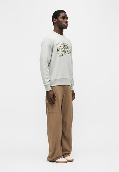 Άνδρας που στέκεται πλαγίως, φοράει ανοιχτό γκρι φούτερ με τυπωμένο κείμενο, φαρδιά καφέ παντελόνια και λευκά παπούτσια μπροστά σε απλό φόντο.