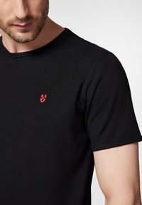 Czarna koszulka z krótkim rękawem wykonana z gładkiego materiału, z małym czerwonym logo na piersi. Klasyczny krój z okrągłym dekoltem, staranne wykończenie.