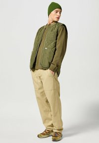Gilet trapuntato verde sopra una camicia a maniche lunghe color khaki, abbinato a pantaloni beige e scarpe marroni con accenti colorati. Cappello di maglia testurizzata indossato.