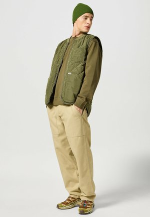Gilet trapuntato verde sopra una camicia a maniche lunghe color khaki, abbinato a pantaloni beige e scarpe marroni con accenti colorati. Cappello di maglia testurizzata indossato.