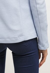 Haut à manches longues bleu clair texturé, associé à un jean slim bleu foncé, montrant une vue de dos et de côté du bas du torse et de la main droite.