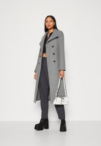 Manteau en laine gris avec un col à revers et une double boutonnage, porté par-dessus un haut court noir et un pantalon gris. Bottes à plateforme noires et sac blanc.