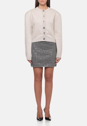 Donna che indossa un cardigan corto beige con bottoni a lettere e una minigonna grigia con motivo, abbinata a tacchi neri a punta.