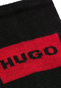 Zwarte sokken met een rode band waarop "HUGO" in vetgedrukte zwarte letters staat. De stof is glad met een gebreide textuur.