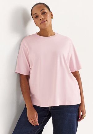 Frau mit goldenen Creolen, die ein hellrosa T-Shirt mit kurzen Ärmeln und dunkelblaue Jeans trägt, steht vor einer weißen Wand.