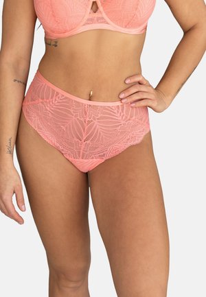Woman wearing high-waisted peach lace lingerie bottom and matching bra, with a hand on her hip and visible tattoos on her arms.