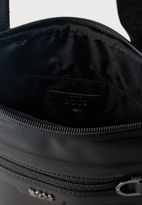 Μαύρη τσάντα από υφασμάτινη υφή με μερικώς ανοιχτό φερμουάρ που αποκαλύπτει μαύρη εσωτερική επένδυση και μια μικρή ετικέτα με ανάγλυφη την επιγραφή "BOSS".