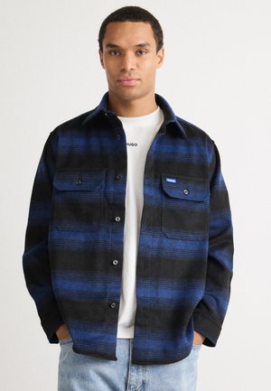 Flanellen shirt in zwart en blauw met horizontale strepen, borstzak, knoopsluitingen, gedragen over een wit T-shirt, gecombineerd met lichte spijkerbroeken.