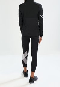 Svart sportjacka med hög krage, matchad med svarta leggings med vita geometriska mönster. Båda är designade för aktivt bruk.