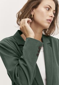 Zaļš žakets ar šauru piegriezumu, ar kļavu apkakli un trim brūniem pogām uz aproces, uzvilkts virs baltā topa ar rakstainu tekstūru.