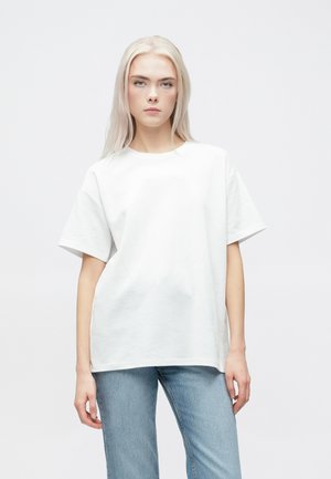 Biała, oversize bawełniana koszulka z krótkim rękawem i okrągłym dekoltem, prezentowana na jednolitym tle, w zestawie z jasnoniebieskimi dżinsami.