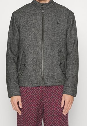 Herringbone-patterned gray jacket with a high collar, front zipper, and side pockets; worn over patterned maroon pants.