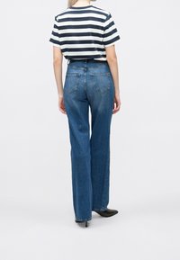 Žena v modrých vysokých rovně střižených džínách, černých lodičkách s špičatou špičkou a bílo-námořnickém pruhovaném tričku s krátkým rukávem, stojí zády.