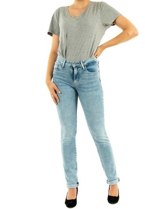 Frau trägt graues T-Shirt mit kurzen Ärmeln, hellblaue hoch taillierte Skinny-Jeans und schwarze Ballerinas, steht mit einer Hand in der Hüfte.