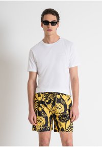 Camiseta blanca de manga corta con un sutil logotipo, combinada con bañadores amarillos que presentan un patrón de hojas tropicales en negro. Las gafas de sol completan el look.