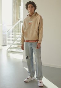 Beige hettegenser med "PUMA"-logo, lagt over en rød tie-dye skjorte. Lyseblå slitte jeans og hvite sneakers med røde detaljer.
