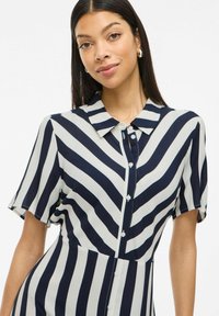 Marinblå och crème randig skjortklänning med krage, korta ärmar och figurnära midja. Slät tyg med en lätt lyster.