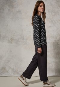 Svartvitt mönstrad långärmad topp, mörkgrå vida byxor och beige sneakers med bruna accenter, mot en neutral bakgrund.
