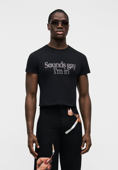 Tricou negru cropped cu text din strasuri „Sounds gay I'm in.” Modelul poartă pantaloni negri cu un design imprimat de mână și țigară.