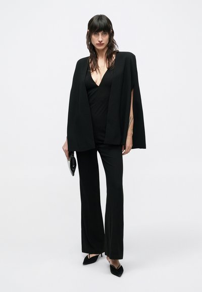 Black jumpsuit with a deep V-neck, complemented by a black cape draped over shoulders. Model holds a small black clutch, wearing black pointed heels.