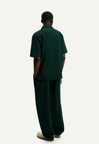 Dark green short-sleeve shirt with a collar, paired with loose-fitting trousers in matching fabric. Features side pockets and textured material.