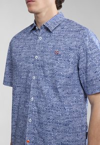 Camisa de botones de manga corta con un patrón azul y blanco. Presenta un cuello estructurado, un logo en el pecho y acentos de botón en contraste.