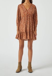 Donna che indossa un mini abito floreale color ruggine con maniche a tre quarti e stivaletti alla caviglia in suede beige, in piedi su uno sfondo chiaro.