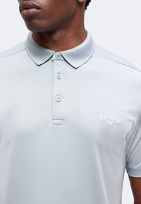Homme portant un polo gris clair avec une patte de trois boutons et un logo Callaway blanc sur la poitrine.
