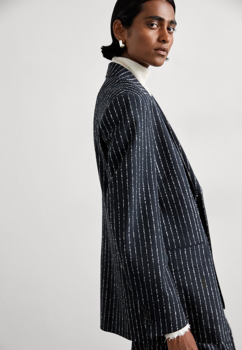 Granatowa marynarka w prążki z białymi, teksturowanymi liniami, o dopasowanym kroju, z klapami typu notch i subtelnymi, postrzępionymi detalami przy mankietach.